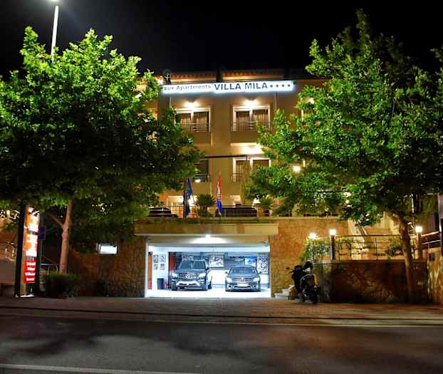 Hotellbygning med opplyst inngang og parkeringsgarasje, omgitt av trær, om natten. (AI-generert tekst)