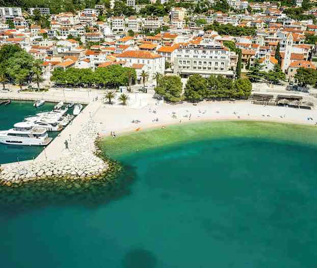 Flyfoto av en strand med turkis vann og en brygge med båter, omgitt av bygninger. (AI-generert tekst)
