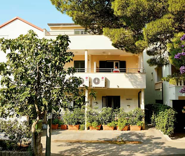 Toetasjes hus med balkonger og klimaanlegg, omgitt av grøntområder og blomster. (AI-generert tekst)