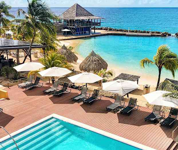 Utsikt over en strand med solsenger, parasoller, palmer og et basseng. Havet er synlig i bakgrunnen. (AI-generert tekst)