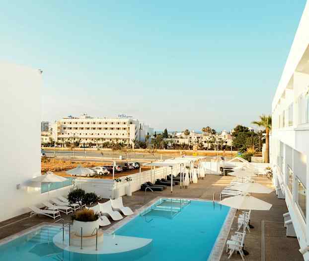 Hotellbasseng med solsenger og parasoller, utsikt mot gaten og omkringliggende bygninger. (AI-generert tekst)