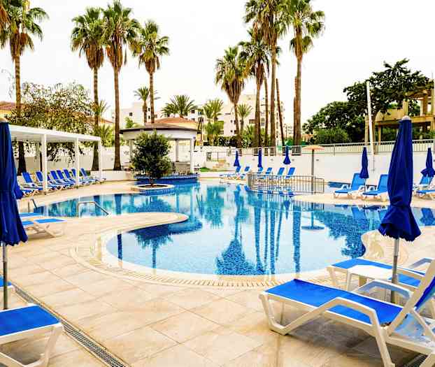 Bilde av et utendørs svømmebasseng omgitt av solsenger, blå parasoller og palmer. (AI-generert tekst)