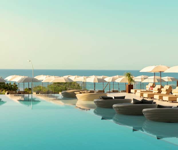 Et infinitybasseng med solsenger og parasoller ved havet. (AI-generert tekst)