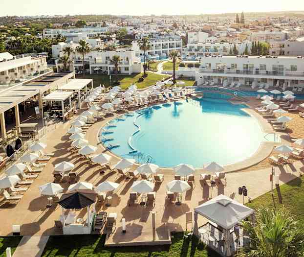 Et stort utendørsbasseng omgitt av solsenger og parasoller, med hotellbygninger i bakgrunnen. (AI-generert tekst)