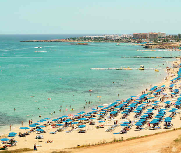 Utsikt over en strand med blå parasoller og badegjester ved det klarblå havet. (AI-generert tekst)