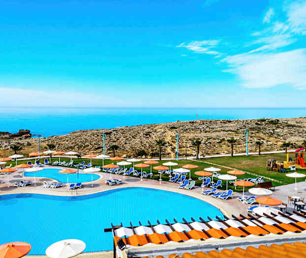 Utsikt over et basseng med solsenger og parasoller, mot havet og steinete landskap. (AI-generert tekst)