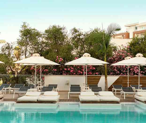 Et bilde av et svømmebasseng med solsenger og parasoller, omgitt av frodig vegetasjon og en lyseblå himmel. (AI-generert tekst)