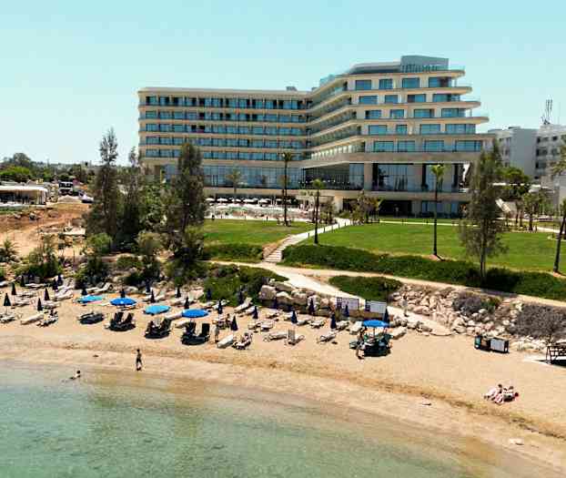 Bilde av en sandstrand med oppstilte parasoller og solsenger foran en moderne hotellbygning og grøntområder. (AI-generert tekst)