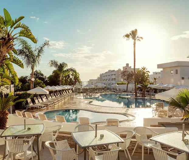 Bildet viser et utendørsbasseng på et hotell, omgitt av solsenger og parasoller. I forgrunnen er det bord og stoler under en solrik himmel. (AI-generert tekst)