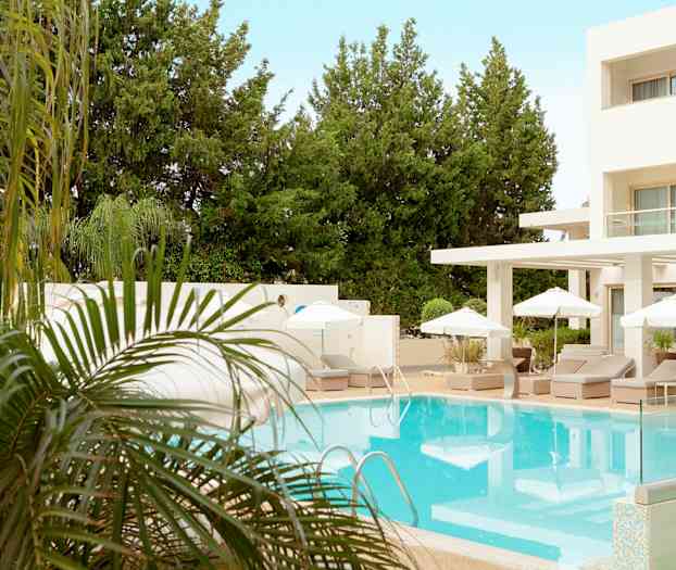 Et utendørs basseng omgitt av solsenger og parasoller ved et hotell med frodige trær i bakgrunnen. (AI-generert tekst)