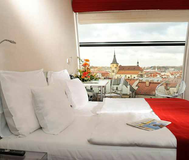 Et hotellrom med en dobbeltseng, dekorert med rødt og hvitt sengetøy. En panoramautsikt over en by med kirkebygg er synlig gjennom vinduet. (AI-generert tekst)
