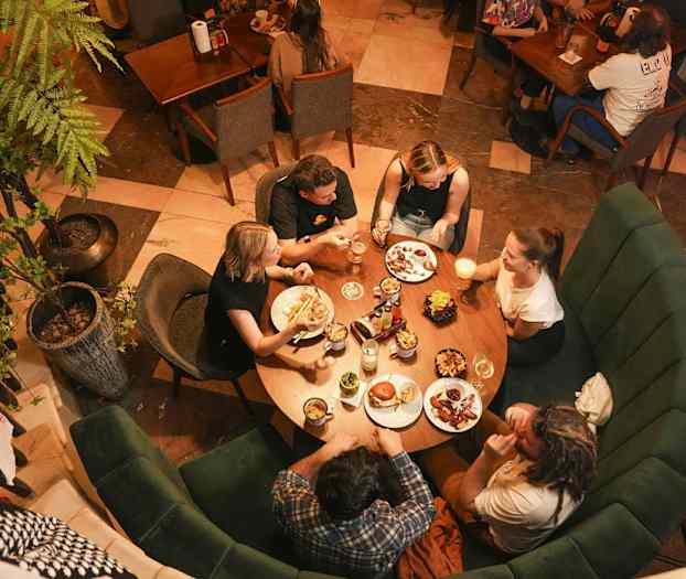 Fem personer sitter rundt et rundt bord på en restaurant og spiser og drikker. (AI-generert tekst)