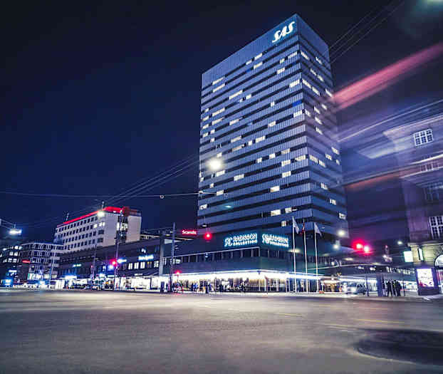 En høy bygning med SAS-logo på toppen, Radisson Collection-hotell på første etasje, omkringliggende bybygninger og nattmiljø. (AI-generert tekst)