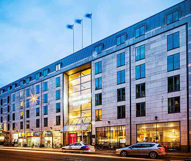 Eksteriør av hotellbygning om kvelden, med belysning og gaten med passerende biler. (AI-generert tekst)