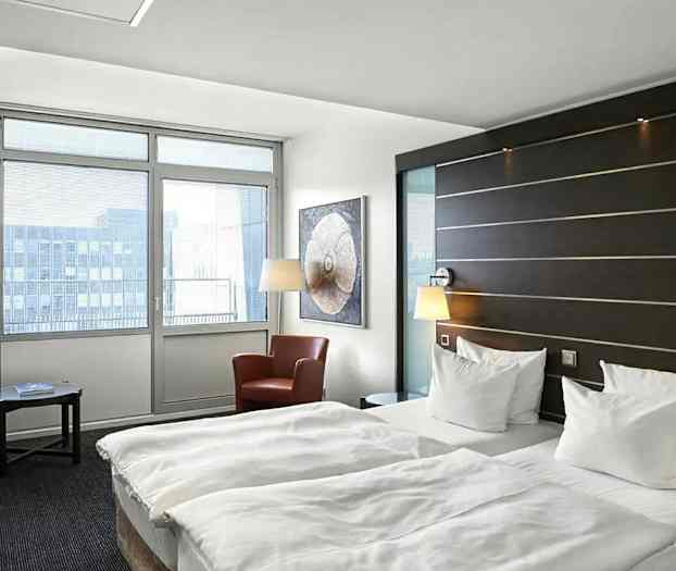 Et hotellrom med to enkeltsenger, en sittegruppe med to lenestoler og et bord samt store vinduer som slipper inn naturlig lys. (AI-generert tekst)