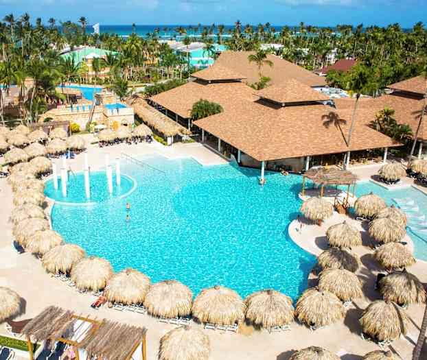 Et hotellområde med et stort basseng omgitt av solsenger med palmeparasoller og bygninger i bakgrunnen. (AI-generert tekst)