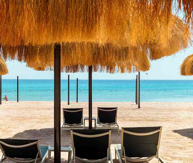 En sandstrand med solsenger og parasoller med halmtak, samt havet i bakgrunnen. (AI-generert tekst)