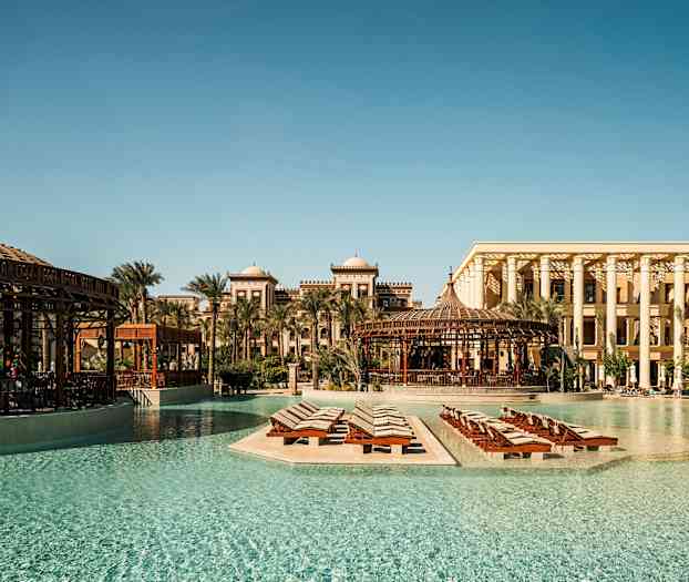 Et postkortlignende utendørsbilde av et luksushotell med basseng og solsenger omgitt av palmer og tradisjonell arkitektur. (AI-generert tekst)