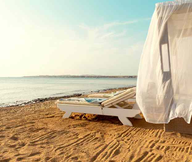 En solseng med et hvitt draperi på en sandstrand ved havet. (AI-generert tekst)