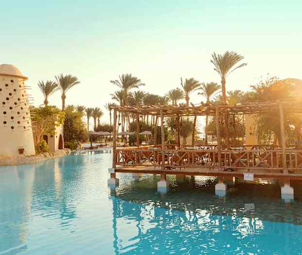 Bilde av et svømmebasseng med en trepaviljong og en bygning med små hull, omgitt av palmer. (AI-generert tekst)