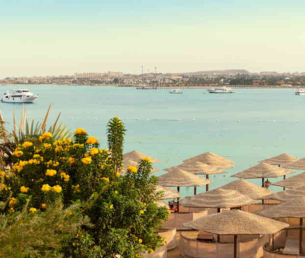 Strand med parasoller, havet og båter i bakgrunnen. (AI-generert tekst)