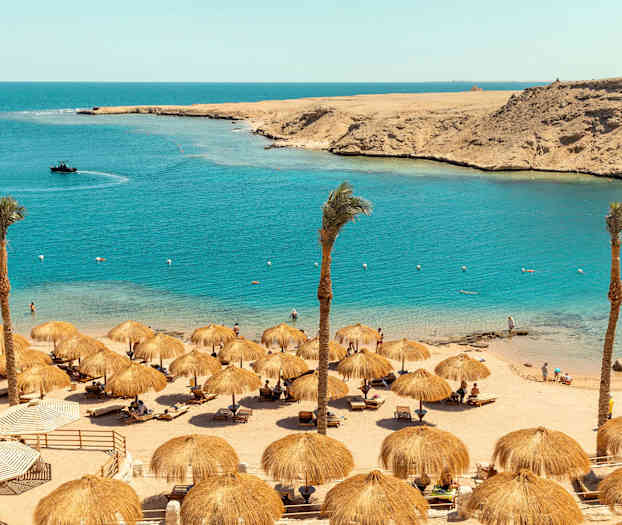 En sandstrand med parasoller, palmer og mennesker ved vannet. (AI-generert tekst)