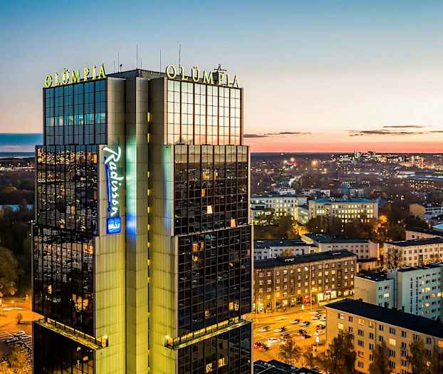 En høy bygning merket "Olümpia" og "Radisson" med byutsikt i skumringen. (AI-generert tekst)