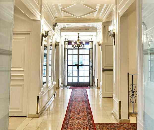 En elegant korridor med hvite vegger og gulv, et rødt teppe og en lysekrone samt glassdører i bakgrunnen. (AI-generert tekst)