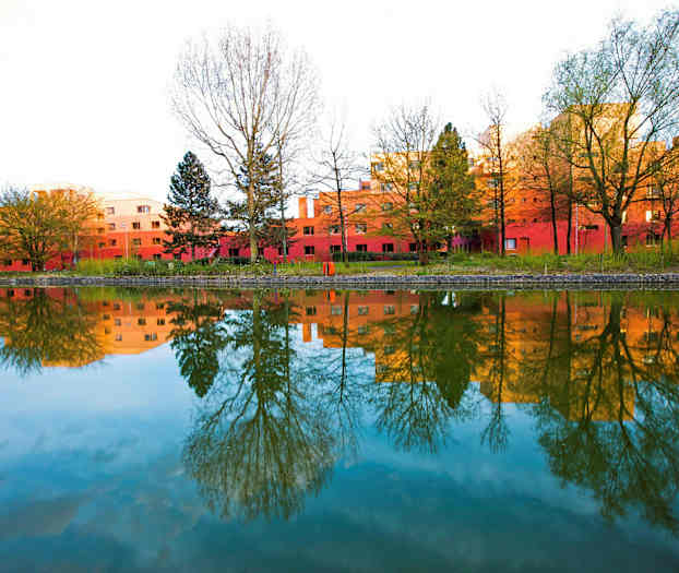 Et bilde av røde bygninger omgitt av trær, som reflekteres i en stille vannoverflate. (AI-generert tekst)