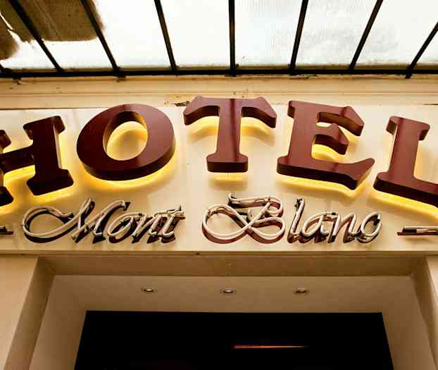 Et hotellskilt med teksten "HOTEL Mont Blanc", der "HOTEL" er i store opplyste bokstaver og "Mont Blanc" er i en kursiv dekorativ stil. (AI-generert tekst)