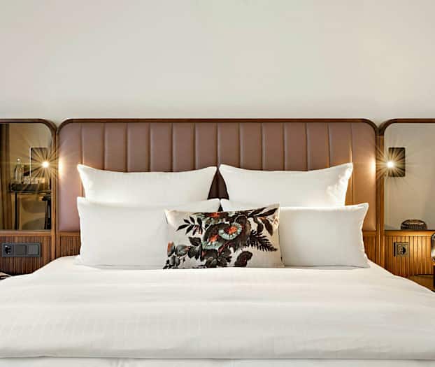 En dobbeltseng med hvitt sengetøy, flere puter og en dekorativ pute med blomsterdesign. Sengens gavl er brun med speil på sidene. (AI-generert tekst)