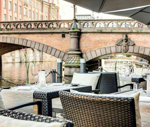 Pent oppsatte bord på en terrasse med utsikt over en bro og en elv. (AI-generert tekst)