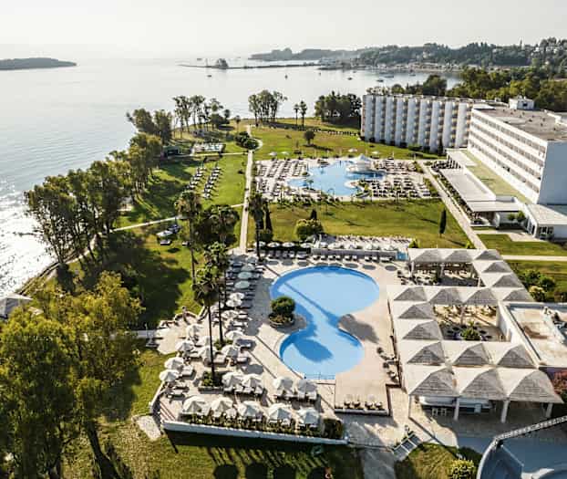 Luftfoto av et hotellområde ved sjøen med bassenger, grøntområder og bygninger. (AI-generert tekst)