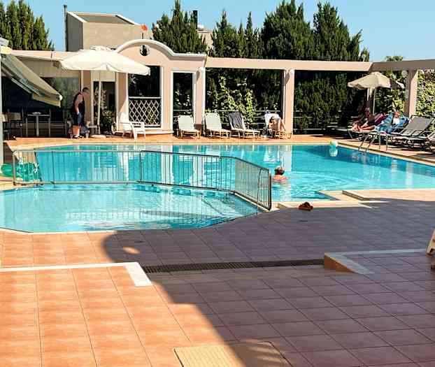 Utendørsbasseng med solsenger og parasoller på siden, samt mennesker som svømmer og soler seg. (AI-generert tekst)