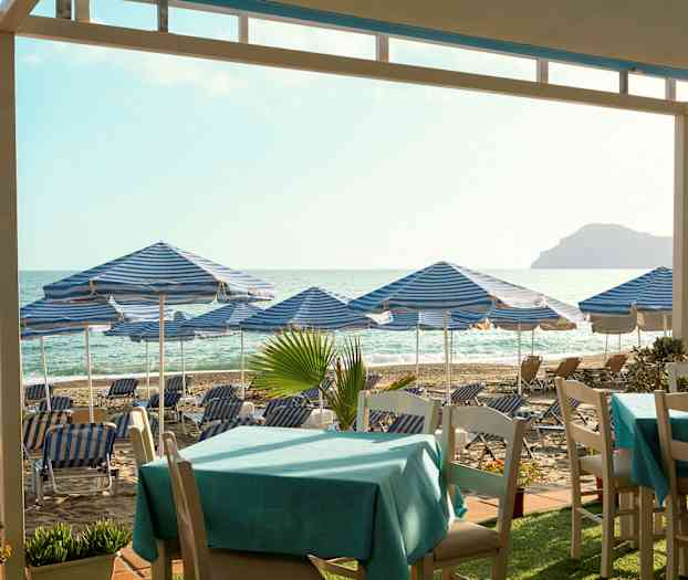 Strand med blå-hvitrutete parasoller og solsenger, bord i forgrunnen. (AI-generert tekst)