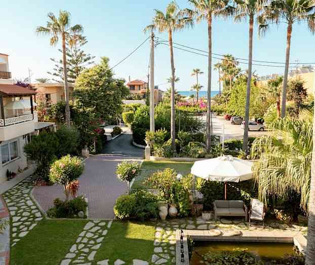 Bilde av en hage med en sofa og parasoll nær en liten dam, omgitt av palmer og grøntområder. (AI-generert tekst)