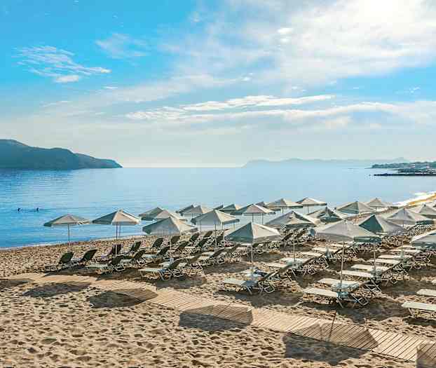 Utsikt over en strand med solsenger og parasoller, omgitt av hav og en fjern øy. (AI-generert tekst)