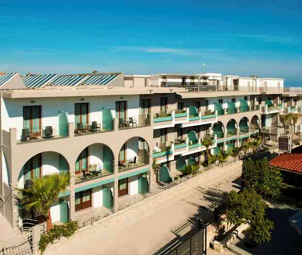 Bilde av et hotell med balkonger og arkitektoniske buer, omgitt av grøntområder. (AI-generert tekst)