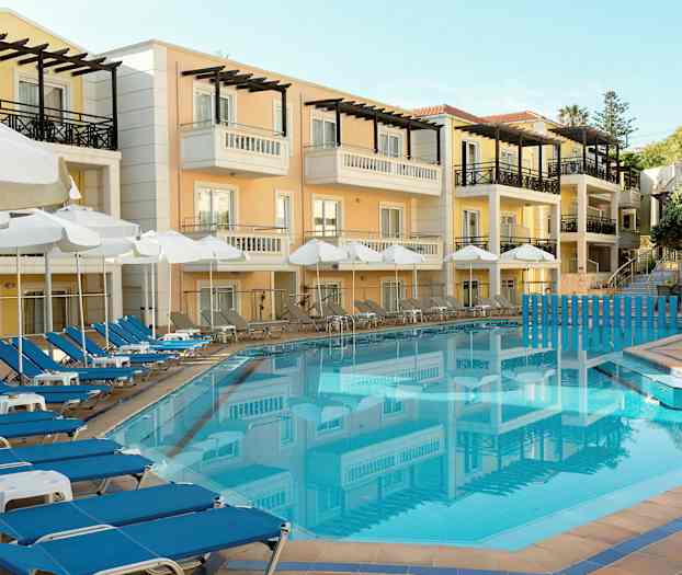 Hotellfasade i pastellfarger med balkonger, foran et blått svømmebasseng omgitt av solsenger og parasoller. (AI-generert tekst)