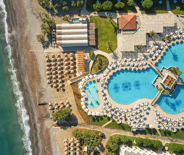 Flyfoto av en strand med parasoller og solsenger ved siden av et stort bassengområde med blå bassenger og hvite parasoller. (AI-generert tekst)