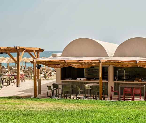 Utendørsrestaurant ved stranden med grønn gressplen, trepergola og parasoller. (AI-generert tekst)