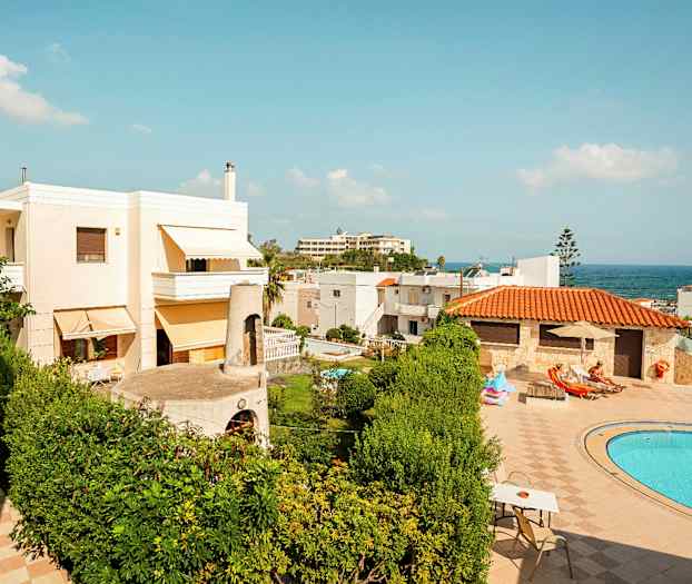 Bildet viser et utendørsområde med hus, grønne planter, et blått svømmebasseng og solsenger på en solrik dag. (AI-generert tekst)