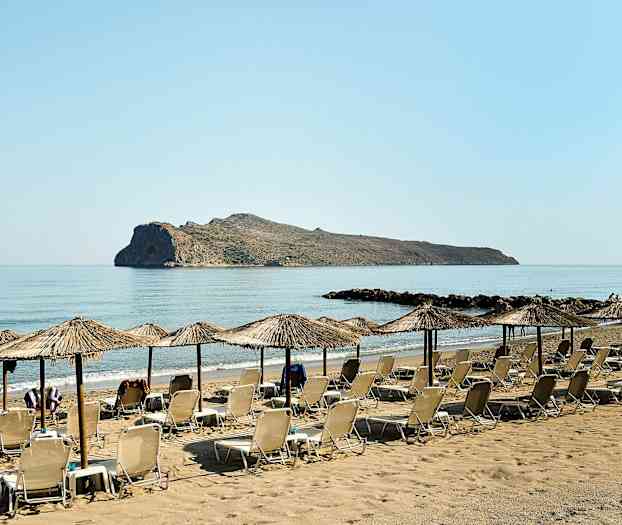 Solstoler og parasoller på en sandstrand med et klippe i havet. (AI-generert tekst)