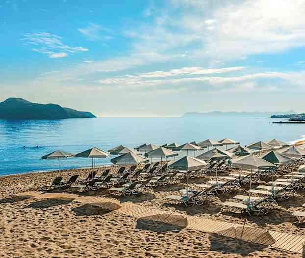 Solstoler og parasoller plassert på en sandstrand ved en rolig havutsikt med en øy og fjell i bakgrunnen. (AI-generert tekst)