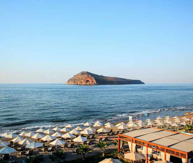 En strand med solsenger og parasoller ved sjøen, med en øy synlig i bakgrunnen. (AI-generert tekst)