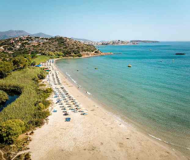 Sandstrand med solsenger ved sjøen, omgitt av grøntområder og fjell i bakgrunnen. (AI-generert tekst)