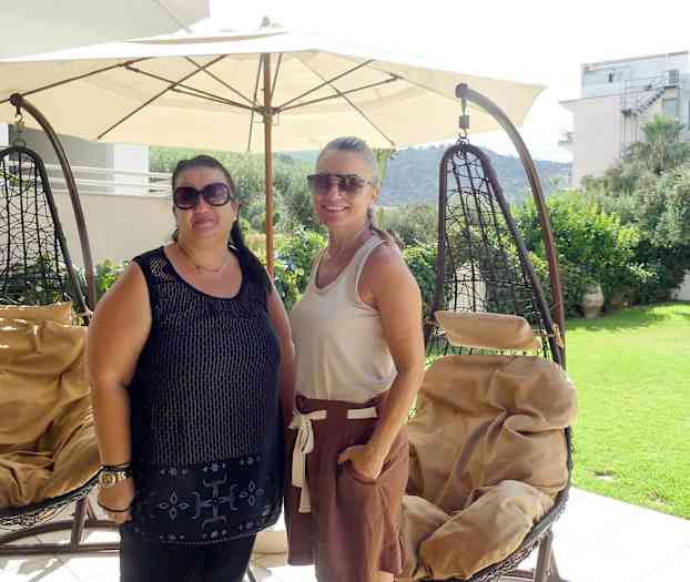 To hengestoler står under en stor parasoll på en terrasse med utsikt over en hage og omkringliggende bygninger. (AI-generert tekst)