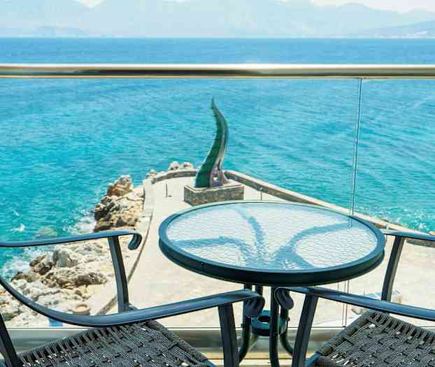 Utsikt fra en terrasse mot sjøen med to stoler og et bord i forgrunnen. (AI-generert tekst)