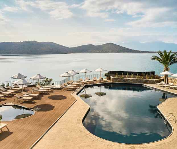 Et basseng med solsenger og parasoller ved sjøen, omgitt av fjell og en palme. (AI-generert tekst)