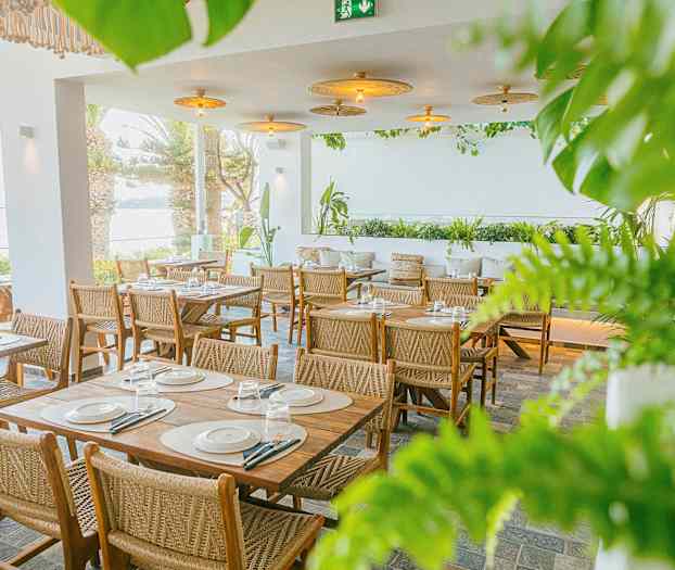 En restaurant med bord og stoler i tre, omgitt av grøntområder og med utsikt over havet. (AI-generert tekst)